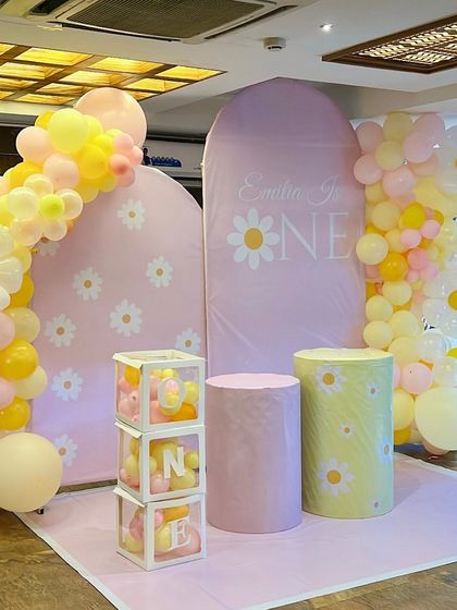 A sweet daisy-themed first birthday party. The decor features daisy-print backdrops, 'ONE' blocks filled with balloons, and a cheerful pink and yellow color palette.
