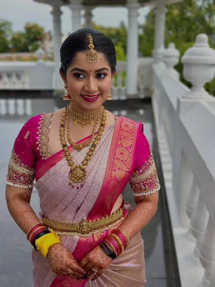 A full view of a stunning bride ready for her ceremony. The vibrant pink and cream saree is paired with classic gold jewelry and a makeup look featuring a bold pink lip and defined eyes, creating a cohesive and regal appearance.