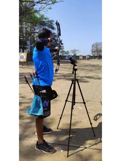 A wide shot of an archer practicing with a compound bow and spotting scope. This setup is typical for serious practice, allowing for immediate feedback on arrow placement.