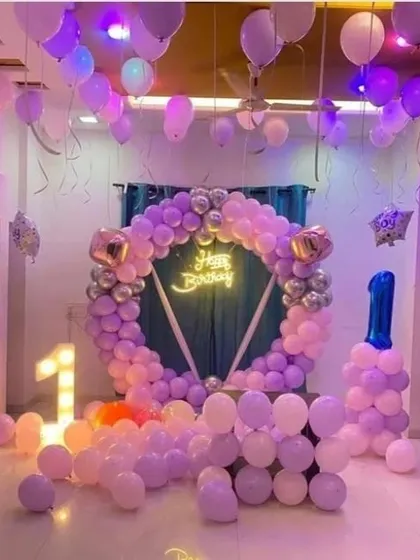 A beautiful first birthday party setup in shades of purple and pink. The room is filled with balloons, featuring a circular arch, a neon sign, and light-up numbers.