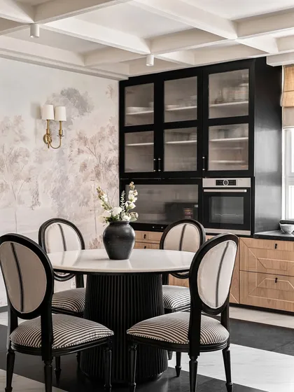 The informal dining area in Riviera Maison, where we paired a fluted-base marble-top table with classic striped dining chairs. The black and oak cabinetry and a soft landscape mural create a sophisticated, European-inspired look.