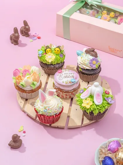 A set of six adorable Easter cupcakes on a wooden platter, decorated with buttercream grass, bunny ears, and tiny chicks. These are a whimsical and delicious addition to your dessert table.