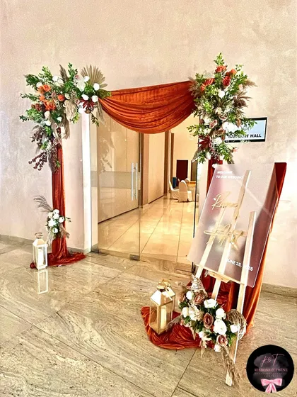 A beautifully styled wedding hall entrance, featuring an asymmetrical arch with rust-colored fabric, pampas grass, and floral arrangements, complemented by a personalized acrylic welcome sign on an easel.