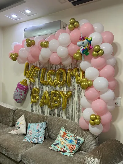 A classic "Welcome Baby" decoration for a living room. This setup features a pink, white, and gold balloon arch over the sofa, a silver foil curtain backdrop, and large gold foil letters.