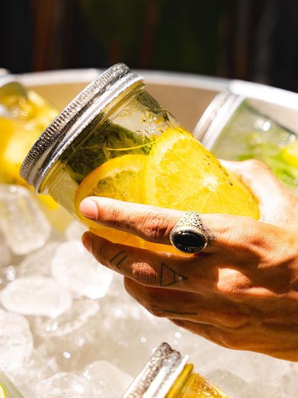 A close-up of a hand grabbing a chilled mason jar beverage, showcasing the fresh fruit and herbs inside.