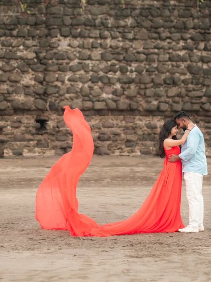 An embrace on the sand. The flowing red trail gown adds a touch of cinematic romance to this beach pre-wedding photo against a historic stone wall.