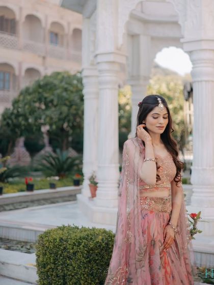 A graceful bridal portrait in a sunlit courtyard, the bride looking down, showcasing the delicate details of her pink Mehendi outfit.