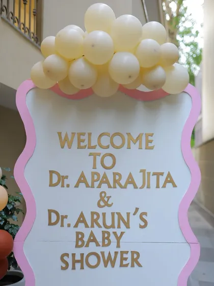A personalized welcome sign for Dr. Aparajita and Dr. Arun's baby shower. The elegant gold lettering and wavy pink border, topped with balloons, create a warm and inviting entrance.