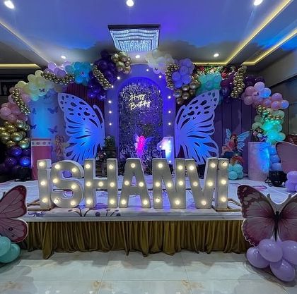 A magical butterfly-themed birthday stage in a banquet hall. The decor features large illuminated butterfly wings, a custom name sign, and a beautiful balloon arch.