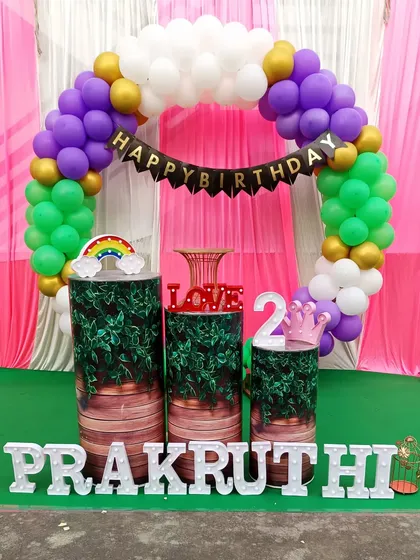 A second birthday party setup with a purple, green, and white balloon arch. The decoration includes themed pedestals and light-up marquee letters on a green carpet.