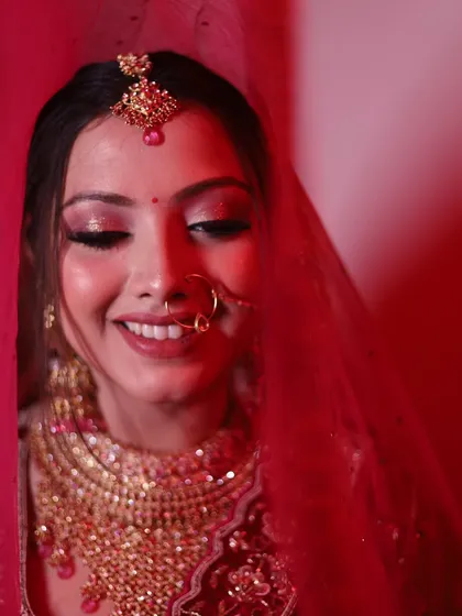 A close-up shot through the red veil, capturing the bride's happy and slightly shy smile. The red hue adds a traditional and vibrant feel.