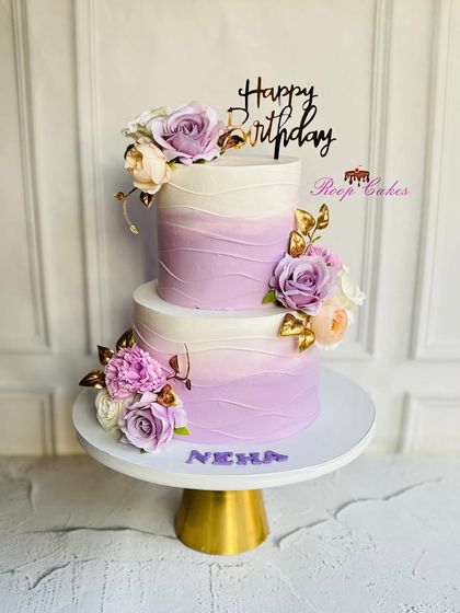 A beautiful two-tier birthday cake with a purple ombre wave design. The cake is decorated with matching purple and white flowers with golden leaves, creating a harmonious and elegant look for Neha's celebration.
