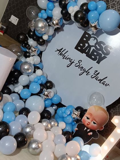 A cool Boss Baby themed party for a first birthday. The decoration uses a custom circular backdrop with the name "Abhiraj Singh Yadav", a balloon cascade in shades of blue, black, and silver, and a light-up number '1'.