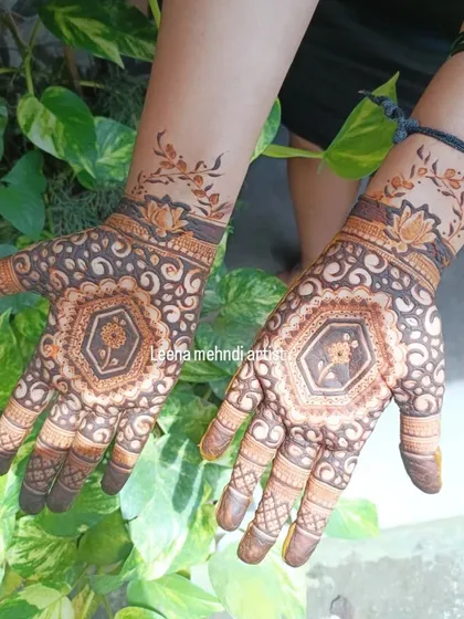 This is an example of modern cut-work, or negative space, mehndi. The design creates a beautiful contrast on the skin, featuring a central floral medallion surrounded by delicate swirls, with a matching vine pattern on the wrist.
