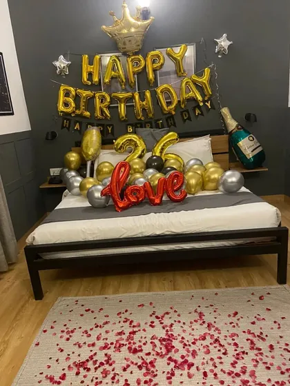 A full view of a stylish and romantic birthday room setup. The decor features gold and silver balloons, 'Love' and 'Happy Birthday' foils, and scattered rose petals.