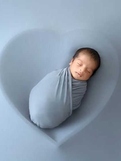 A minimalist portrait of a newborn swaddled in grey, peacefully asleep in a matching heart-shaped bowl.