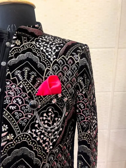 A close-up of a black velvet Indo-Western jacket with intricate multi-colored embroidery. The pink pocket square adds a pop of color.