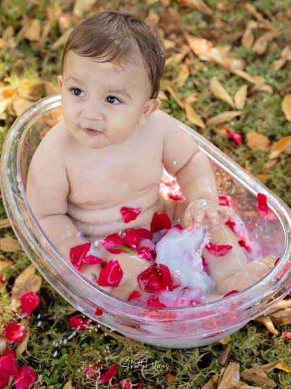 A baby boy enjoys a milk bath with red rose petals in a beautiful outdoor setting.