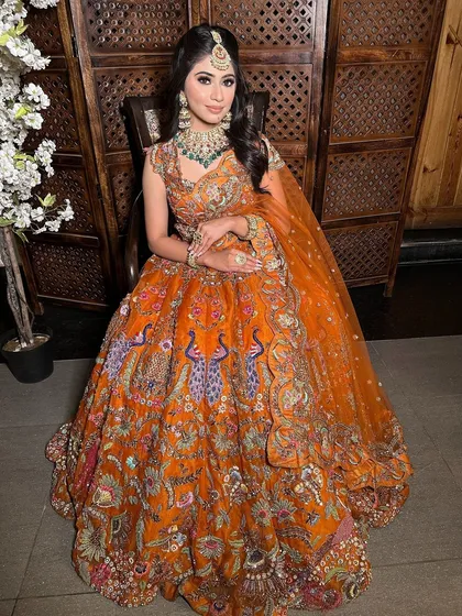 A full-length view of our client's stunning orange engagement lehenga with intricate peacock embroidery. Her subtle makeup and elegant hairstyle complete the look perfectly.
