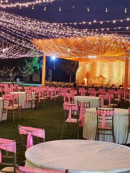 An outdoor reception setting under a canopy of fairy lights. The stage is bathed in a warm golden glow, and the guest tables are decorated with pink accents.
