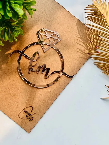The "KM" ring topper styled on its backing board, showing the combination of cursive letters and a modern ring shape.