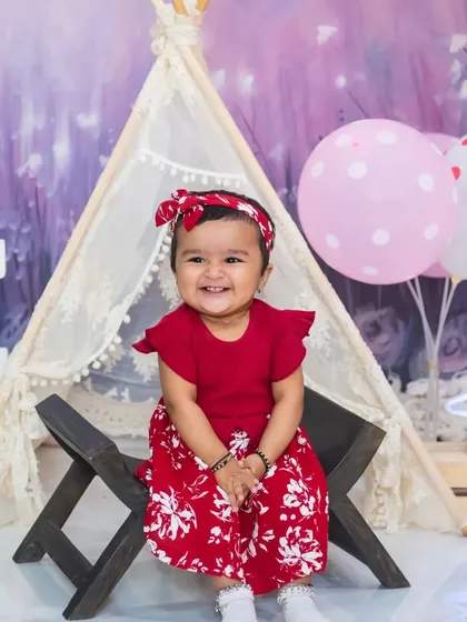 A sweet baby girl in a red dress gives a beautiful, happy smile during her indoor pre-birthday photoshoot.