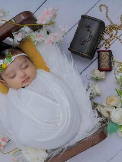 This angelic newborn is swaddled in white and rests in a miniature wooden sleigh. The soft lighting and surrounding white flowers give this portrait a dreamy and ethereal quality.