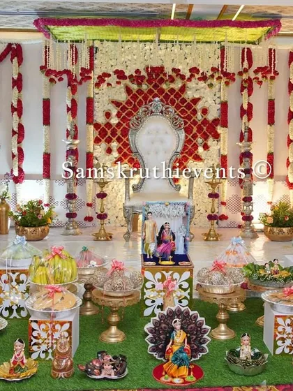 A regal red and white themed baby shower decoration. The backdrop features intricate patterns, complemented by a grand white throne and a symmetrical arrangement of offerings on a green lawn carpet.