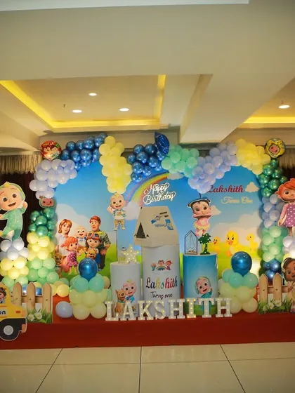 A large-scale Cocomelon theme decoration for a party hall, with a full stage setup including a backdrop, character props, and balloon pillars.