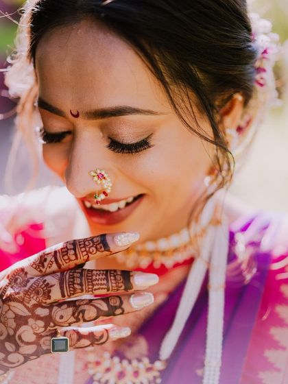 A joyful, sun-kissed portrait of a bride. Her smile is radiant as she looks down at her mehendi-adorned hands, with the light catching the details of her traditional nose ring.