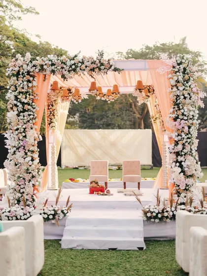 A beautiful pastel-themed mandap, with pillars heavily adorned with white and pink roses. The soft peach draping and hanging floral chandeliers create a dreamy and romantic atmosphere for an outdoor ceremony.