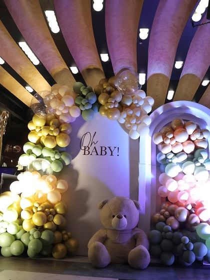 A beautiful 'Oh Baby' photo backdrop with a soft green, gold, and cream balloon arch. The setup is made even cuter with large and small teddy bear props.