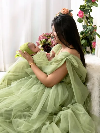 A side profile of a mother in a flowing green dress, cradling her baby. The soft lighting and floral background create a serene and elegant atmosphere.