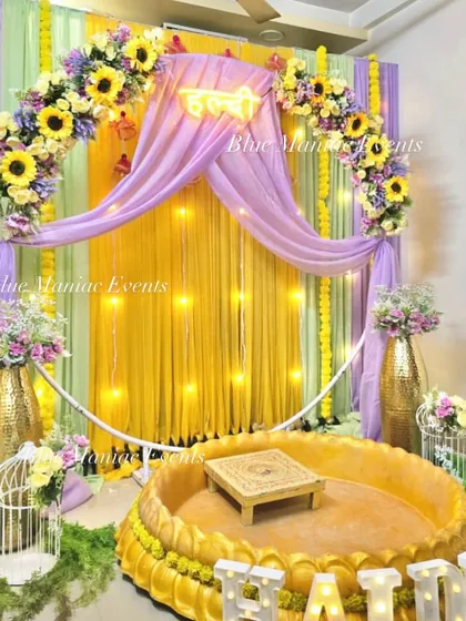 I design classic Haldi ceremony spaces perfect for home functions. This setup features a beautiful floral arch with sunflowers over a yellow and lavender draped backdrop, with a traditional brass-style urli for seating and fun props to get the party started.