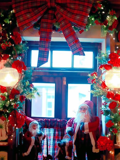 A festive window display with Santa figurines, red flowers, and a large tartan bow, creating a classic Christmas scene.