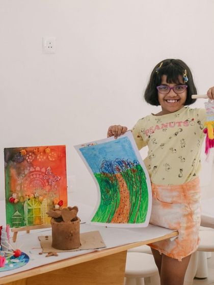 This camper is all smiles as she shows off her art camp creations. She's holding a colorful drawing and a small tapestry, with her other mixed-media projects displayed on the table.