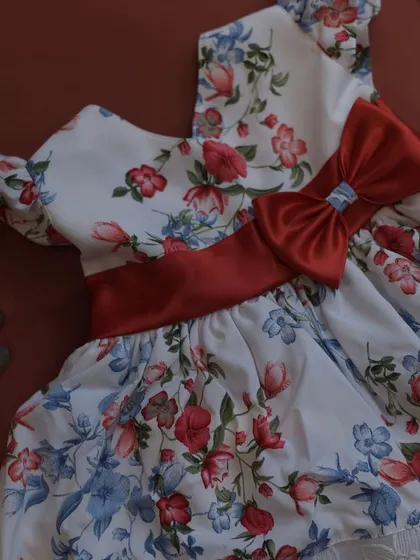 This dress features a beautiful floral print in shades of red and blue on a white background. The design is completed with a rich red satin sash and bow.