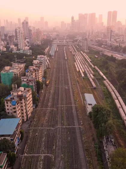 An aerial photograph looking down the railway tracks leading into Mumbai, with trains parked and the hazy city skyline in the distance.