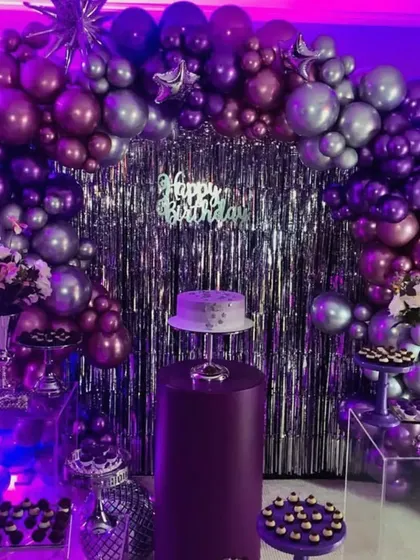 A dramatic and moody party theme with a purple and silver color scheme. The metallic balloons, silver fringe backdrop, and purple uplighting create a sophisticated and celebratory vibe perfect for a milestone birthday.