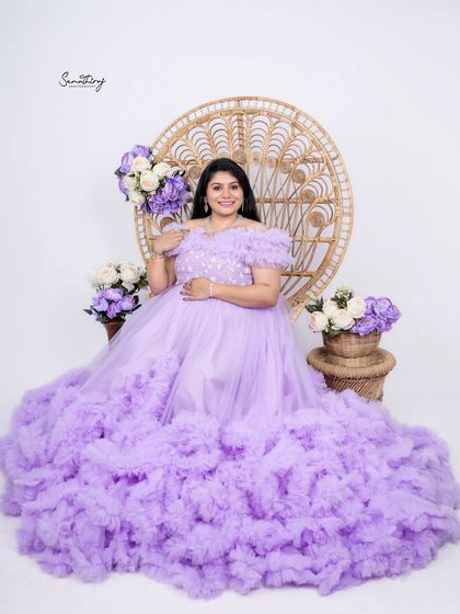 A regal portrait of a mother-to-be in a lavender ruffled gown, seated on a peacock chair. The matching floral arrangements and her confident pose create a stunning and elegant image.