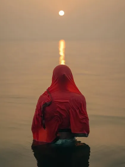 A woman takes a holy dip in the Ganges at sunrise, her red saree a stark contrast against the calm, golden water. This is a moment of pure faith, a quiet prayer in the heart of Varanasi's spiritual landscape.