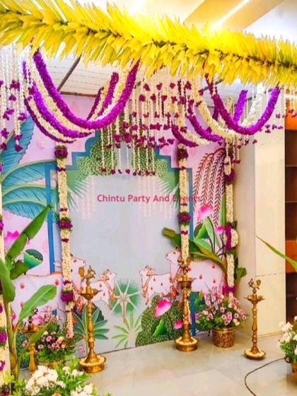 A traditional Seemantham decoration with a Pichwai-style cow art backdrop. The setup is adorned with flower garlands, banana plants, and brass lamps.