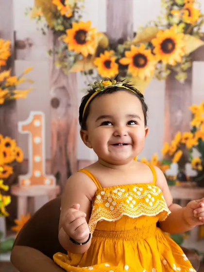 A baby girl celebrates her first birthday with a huge smile, set against a vibrant sunflower-themed backdrop. This is an example of a sitter session, perfect for capturing personality-filled milestones.