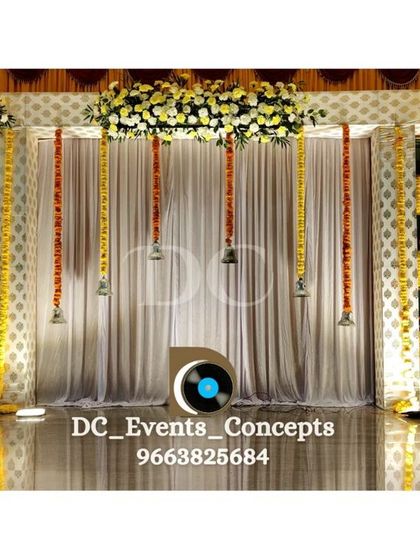 A traditional yet stylish baby shower decor. The backdrop features hanging temple bells and marigold garlands, combining sacred elements with a clean, contemporary presentation.