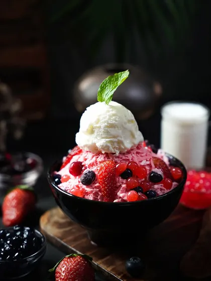 A berry-flavored bingsu or shaved ice dessert topped with vanilla ice cream and a mint leaf. The dark, moody lighting enhances the vibrant red of the berries and the cold, frosty texture.