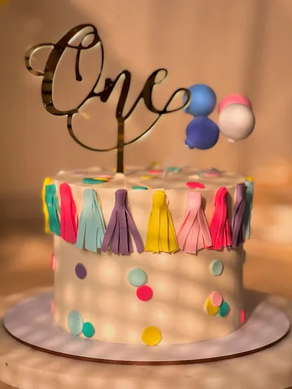 A festive and colorful cake for a first birthday party. This lemon-flavored cake is decorated with a fun tassel garland, confetti dots, and a balloon topper.