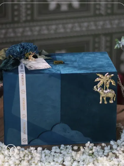 This Rajwada-style hamper features a deep teal velvet chest, perfect for storing keepsakes long after the celebration. The chest is embellished with a golden elephant and palm tree motif, reflecting a blend of Indian heritage and opulent design.