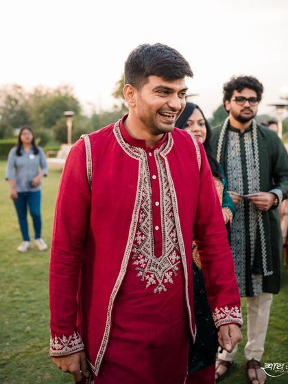 The groom in a handsome red kurta with an embroidered jacket, a perfect look for a festive outdoor celebration.