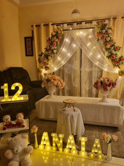 A cozy and romantic 12th birthday or anniversary setup at home, featuring a floral arch with fairy lights and marquee letters.