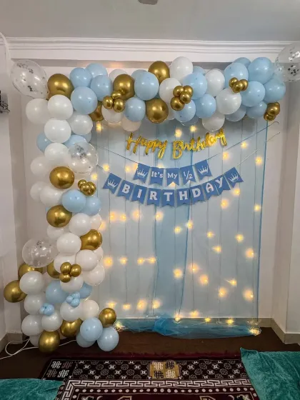 A beautiful half-birthday celebration at home, featuring a blue and gold balloon arch against a curtain backdrop with fairy lights.